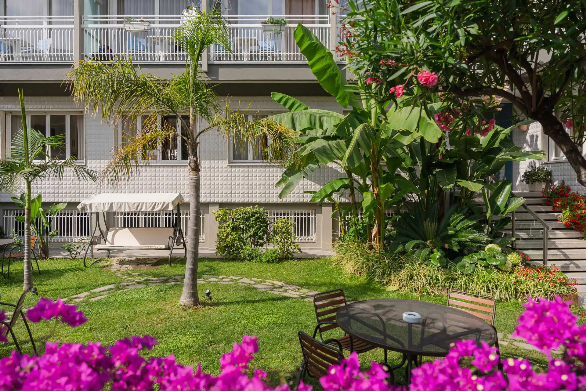 Giardino con tavolo, fiori colorati e piante tropicali davanti a un edificio.