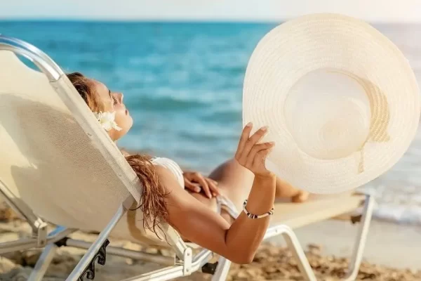 Donna su una sdraio in spiaggia con cappello di paglia e mare sullo sfondo.